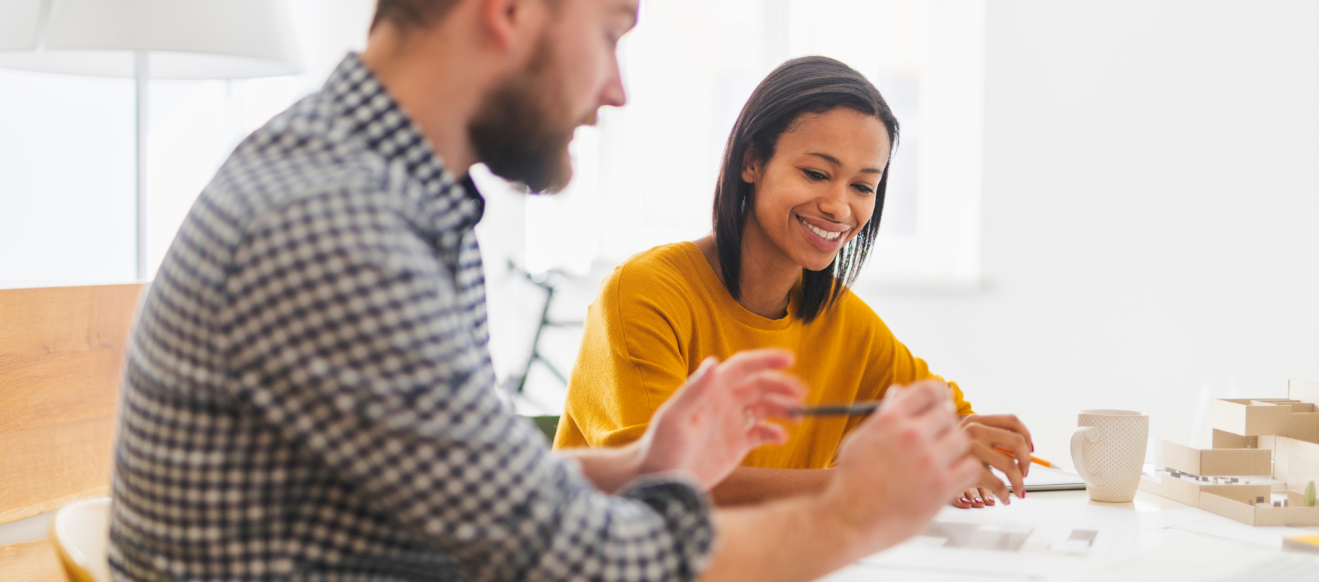 Two people sit at a table working together. The woman in a yellow sweater smiles while holding a pencil. The man in a checkered shirt gestures with a pen as they discuss ideas inspired by CMB Insights. Papers, a mug, and a model are on the table, reflecting their collaborative spirit in this Career Incubator session.