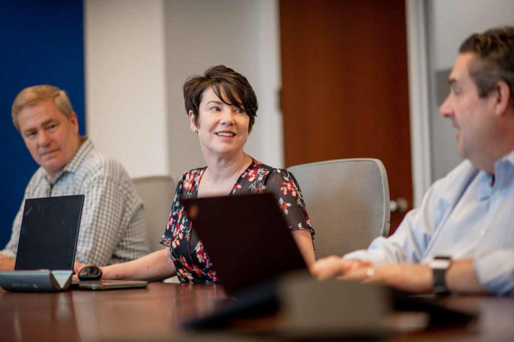 Three people sit at a conference table with laptops, engaged in conversation. The woman in the center is smiling, while the two men on either side listen attentively. The setting appears to be a modern office meeting room.