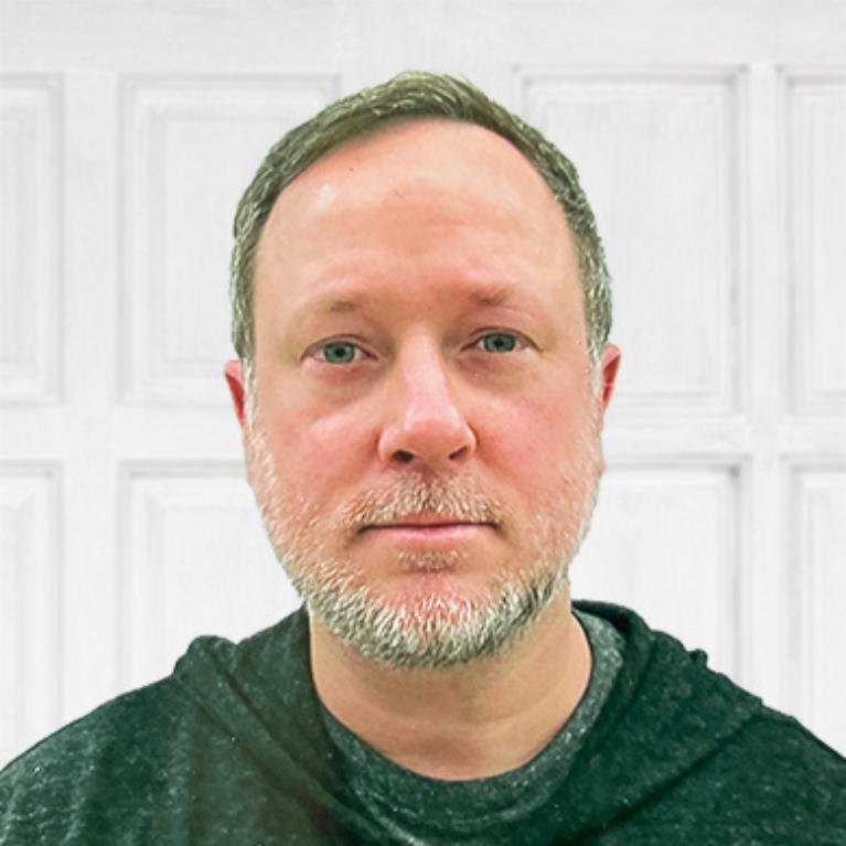 A man with short light brown hair and a beard wears a dark hoodie and stands in front of a white paneled background, looking directly at the camera with a neutral expression.